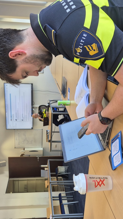 Een student zit achter zijn laptop in een klaslokaal met zijn telefoon in zijn hand