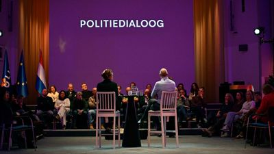 Twee sprekers aan een hoge tafel voor een gevulde zaal tijdens de Politiedialoog