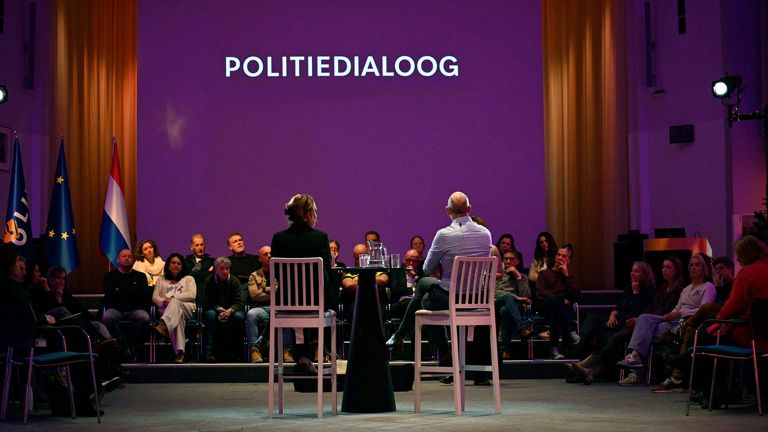 Twee sprekers aan een hoge tafel voor een gevulde zaal tijdens de Politiedialoog