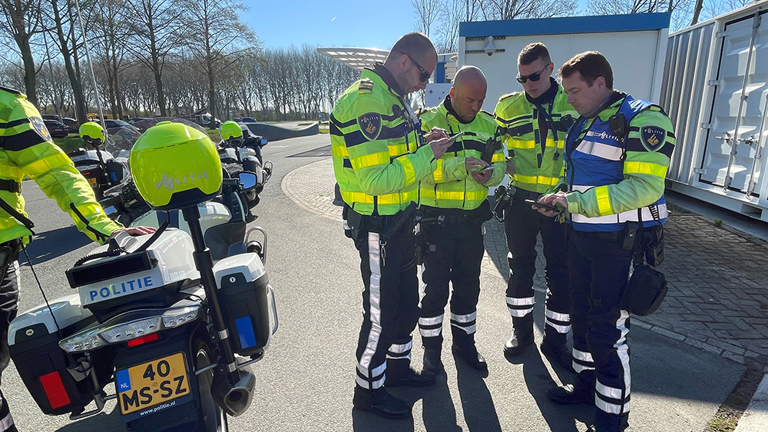 Drie studenten en een docent kijken op hun telefoon naar de route met politiemotoren ernaast