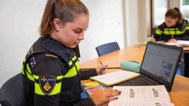 A student in a police uniform sits behind the laptop and looks at a study book