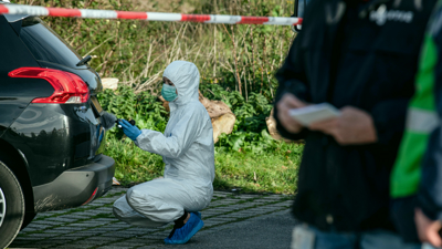 Forensische Opsporing - een vrouw in wit pak zit op haar hurken en doet onderzoek bij de achterkant van een auto